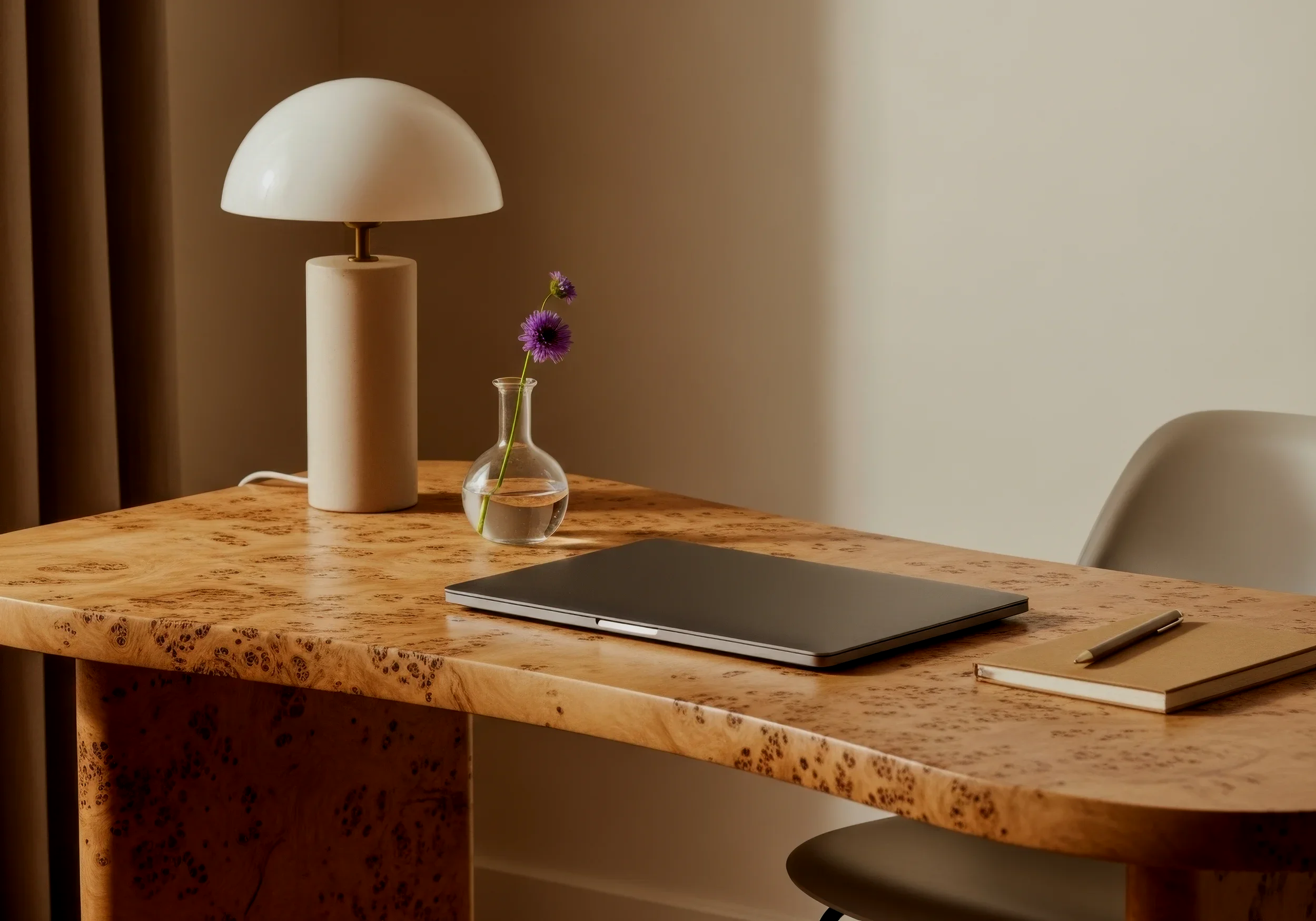 A wooden desk with a closed laptop, a notebook, a pen, a modern table lamp, and a small glass vase with a purple flower.