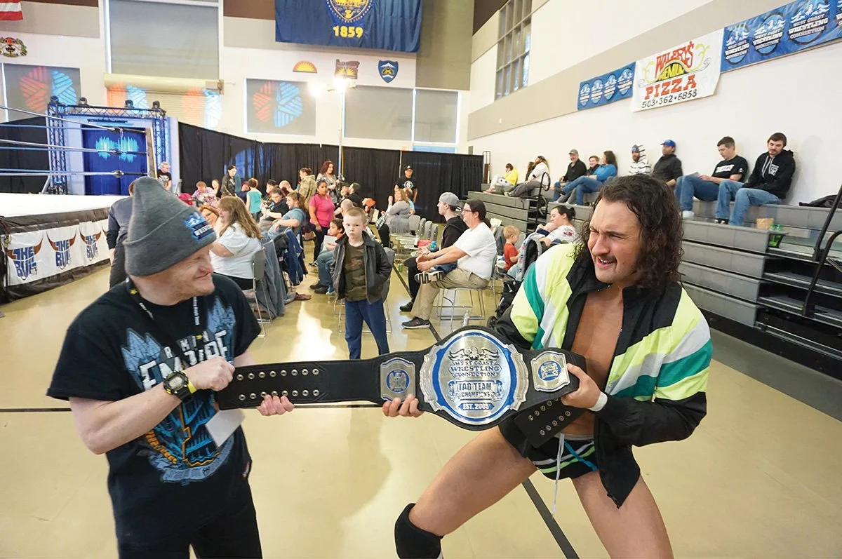 A wrestling event with an audience, a wrestling ring, and two men holding a championship belt. One man is wearing a gray beanie and a black T-shirt, and the other man is dressed in wrestling gear with a green, yellow, and black jacket. The man in wre