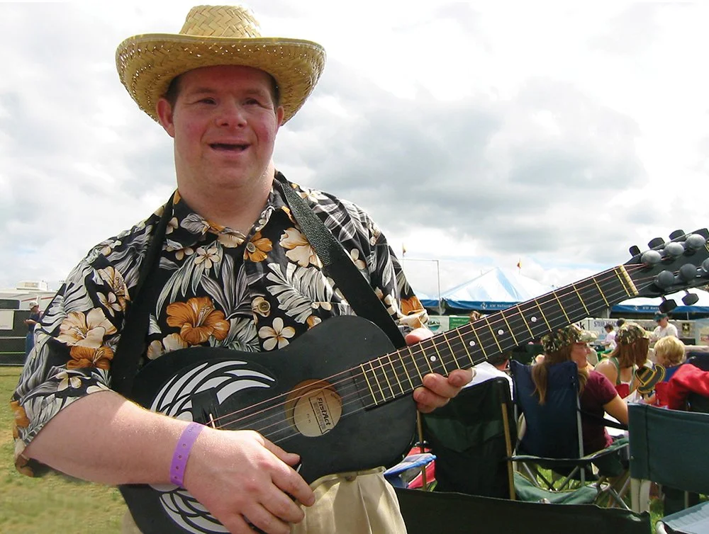 A man wearing a straw hat and a floral shirt holds a black acoustic guitar at an outdoor event with other people seated in tents and chairs in the background.