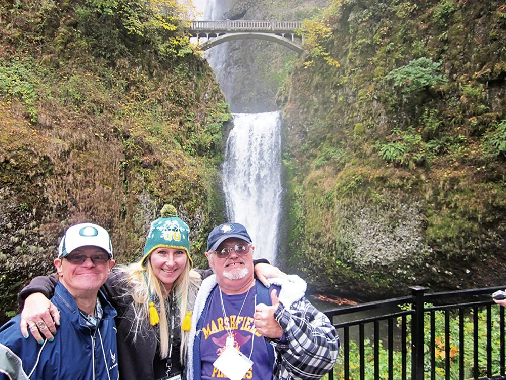 Three people stand in front of a waterfall with a bridge overhead, surrounded by lush greenery.