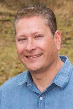 A smiling man wearing a blue denim shirt outdoors with a grassy background.