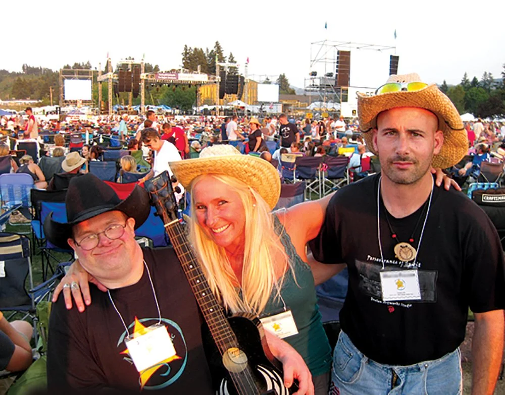 Three people at an outdoor concert, two men and a woman, smiling and wearing straw hats, with a crowd and stage in the background.