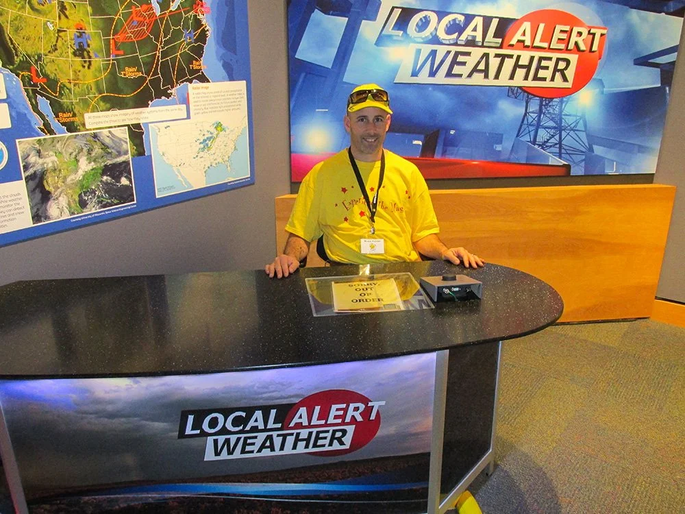 A man sitting at a weather news desk with a sign that reads 'Local Alert Weather', wearing a bright yellow t-shirt and yellow sunglasses, in a weather station with maps and weather graphics behind him.