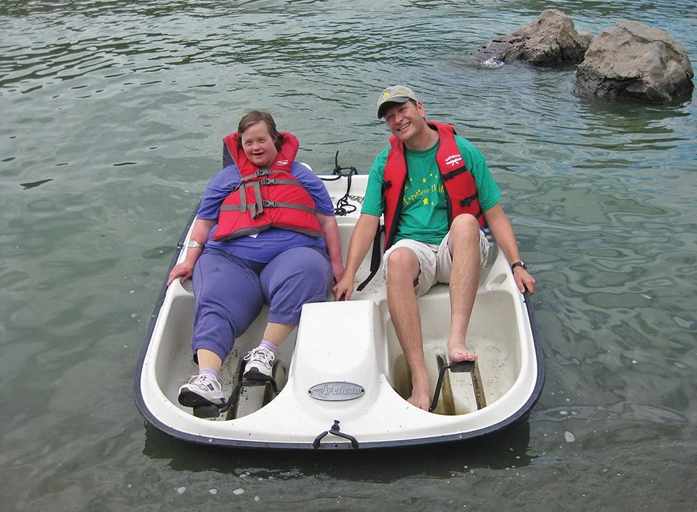 Two people sitting in a small sailboat on the water, holding hands, wearing life jackets, with rocks in the background.