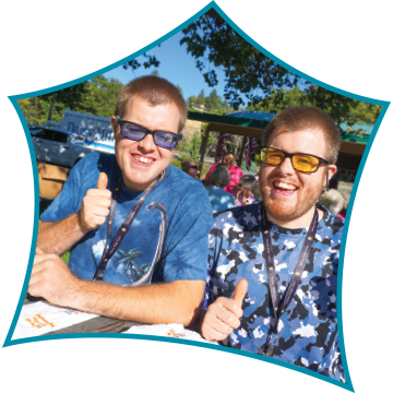 Two smiling young men with glasses giving thumbs up at outdoor event, one in a blue tie-dye shirt, the other in a blue camouflage shirt.