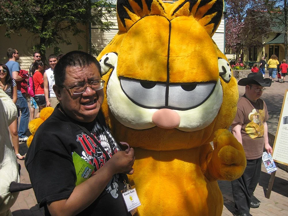A man standing next to a Garfield mascot costume at an outdoor event, with other people in the background.
