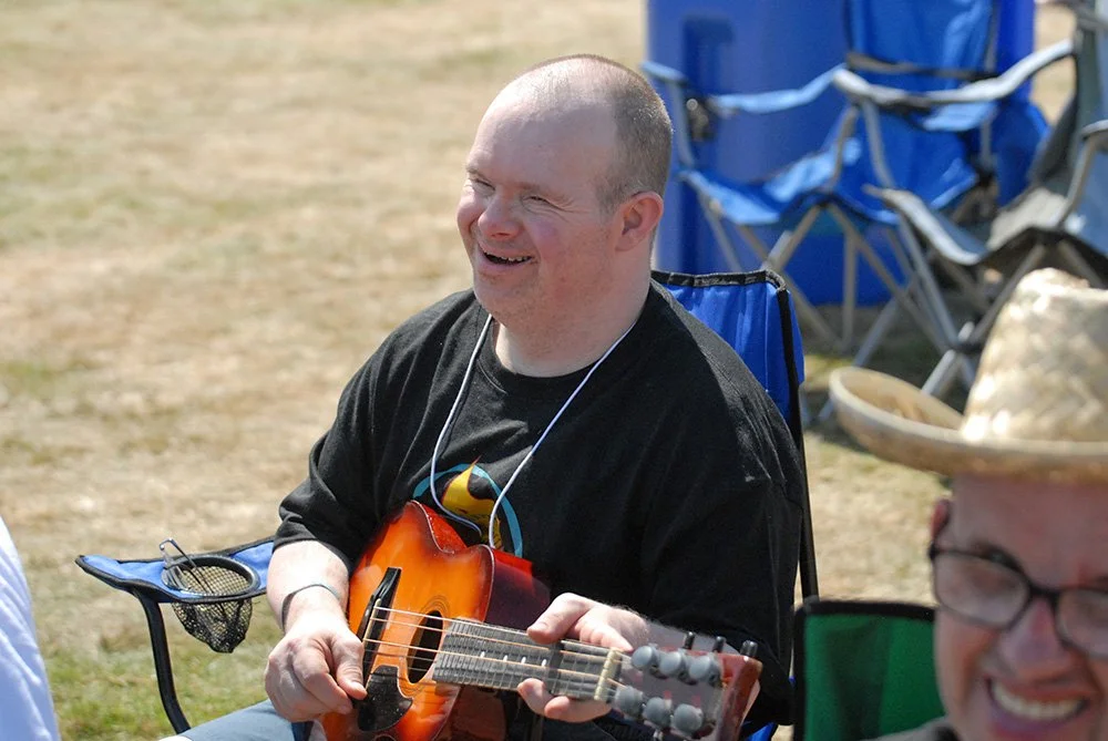 A man smiling and playing a small acoustic guitar, sitting outside at a gathering with outdoor chairs and a tent or canopy in the background.