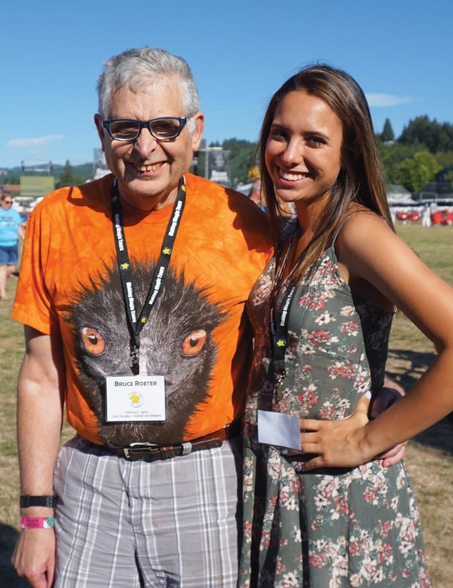 A man and a woman smiling at an outdoor event on a sunny day. The man is wearing glasses, a bright orange T-shirt with a large owl face graphic, and plaid shorts. The woman is wearing a floral dress, has long brown hair, and has her hand on her hip.