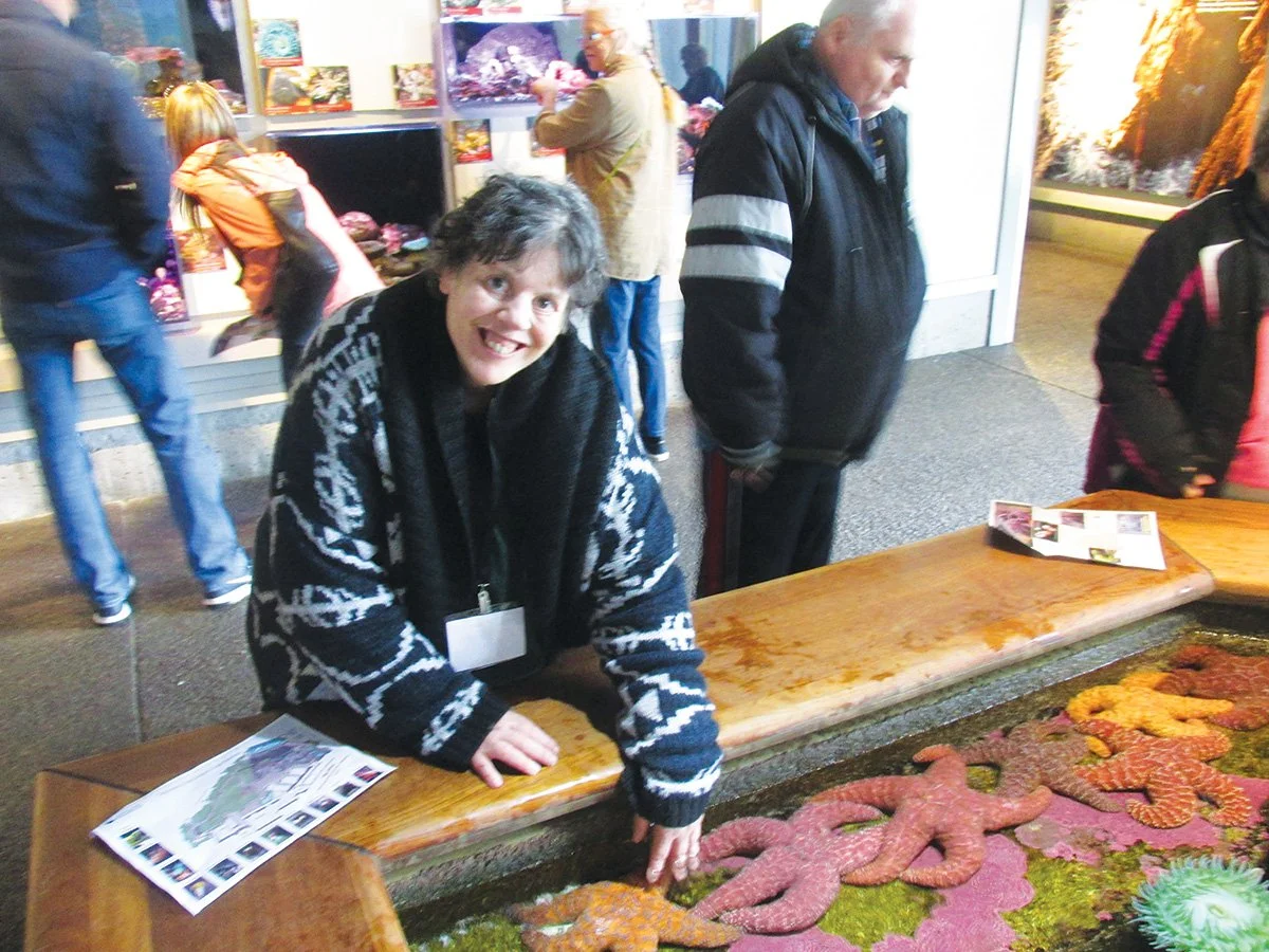 Woman with short dark hair and a black and white sweater smiling and leaning on a display of colorful starfish at an indoor exhibit.