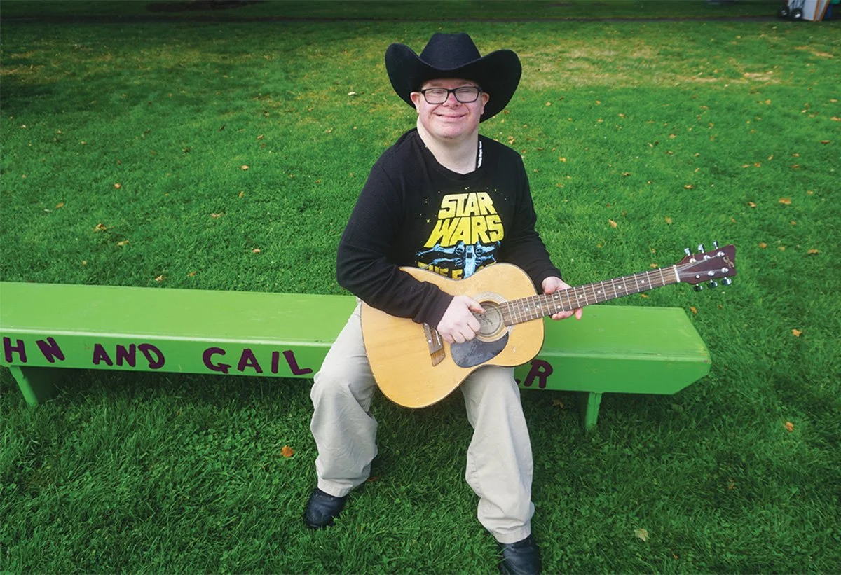 A man wearing a black cowboy hat, glasses, a Star Wars shirt, and beige pants is sitting on a green bench outdoors, holding an acoustic guitar and smiling.