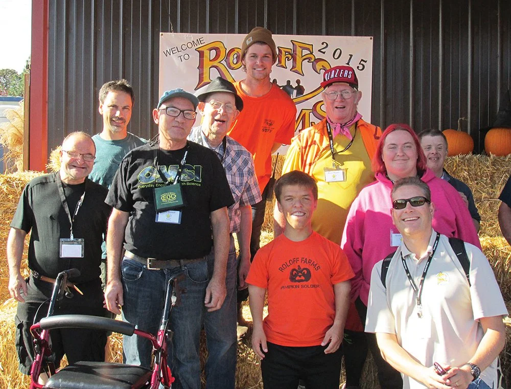 Group of people standing in front of a Halloween pumpkin and hay bales at a fall festival event.