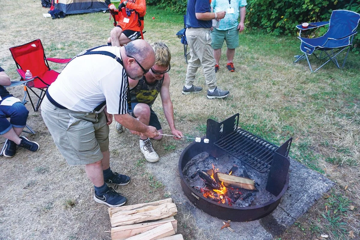 People gathered outdoors at a campsite, starting a fire in a round metal fire pit with logs burning. Some are standing, others sitting in camping chairs, with tents and trees in the background.