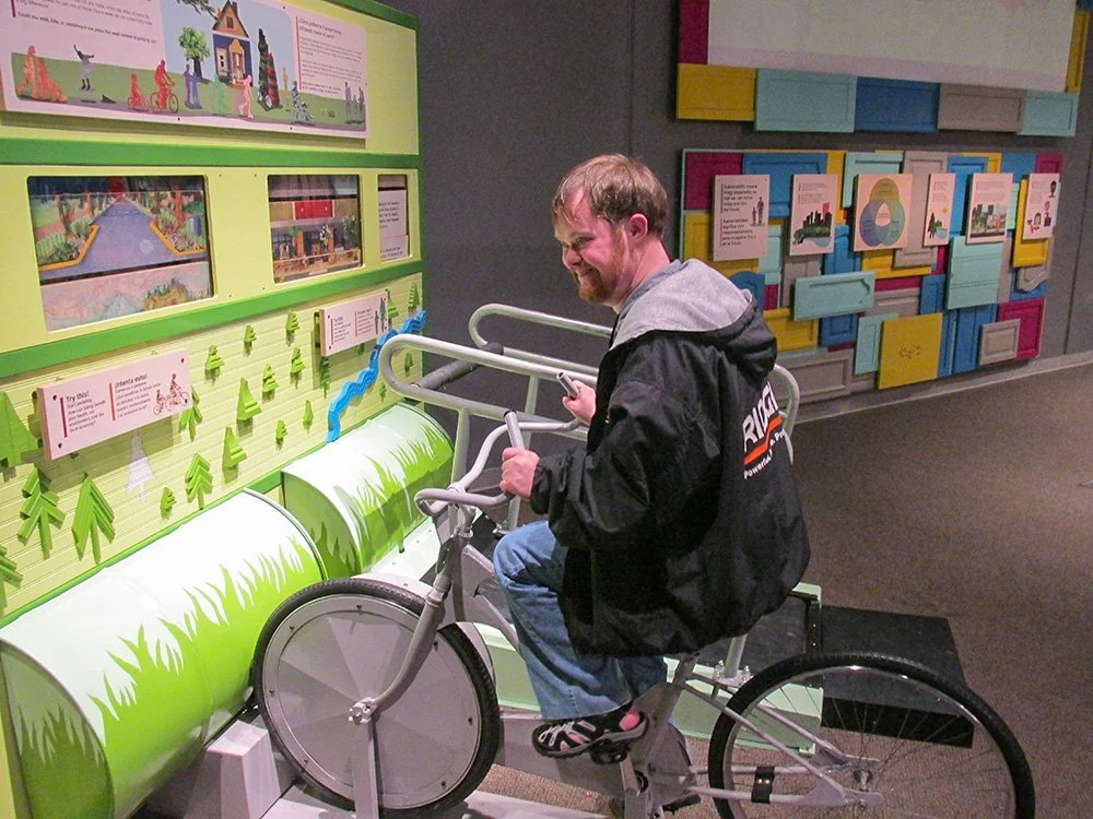 A man riding a stationary bicycle display at a museum or exhibit, with instructional panels and colorful artwork on the walls.