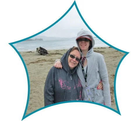 Two women smiling and hugging on a beach with sand, water, and rocks in the background, one wearing sunglasses and a hoodie, the other wearing a hat and sunglasses, framed by a decorative blue outline.