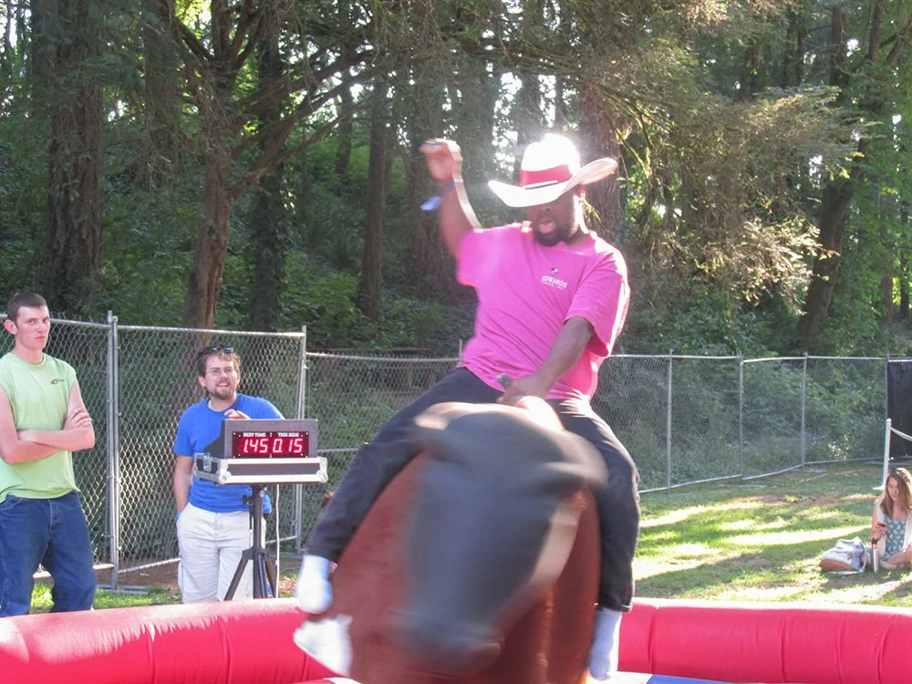 A man wearing a pink shirt and a large cowboy hat rides a mechanical bull at an outdoor event, with two men watching nearby and a woman sitting on the grass in the background.