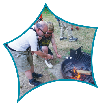 People gathered around a small outdoor fire pit, tending to the fire with logs on a grassy area.