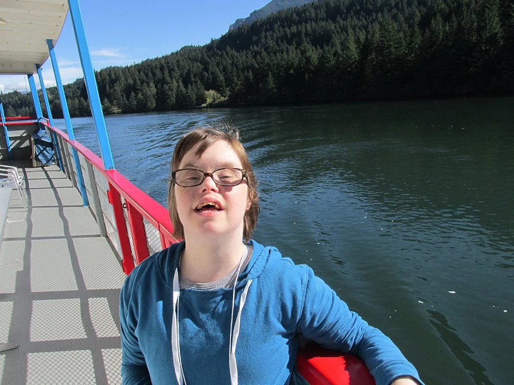 A young person with glasses on a boat, smiling, with a river and forested mountains in the background on a sunny day.