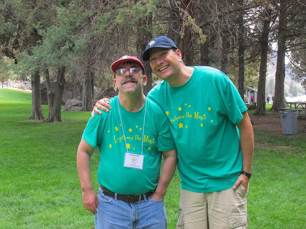 Two men standing outdoors on green grass, smiling. Both are wearing teal T-shirts with yellow stars and the words "Experience the Magic". One man has a red cap and sunglasses, the other has a navy cap. There are trees and park benches in the backgrou
