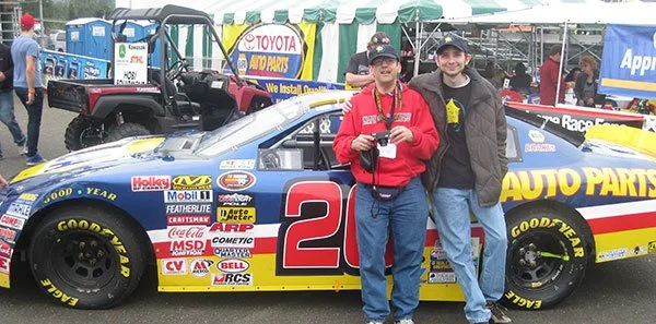 Two men standing in front of a racing car at a motorsports event, with tents and banners in the background.