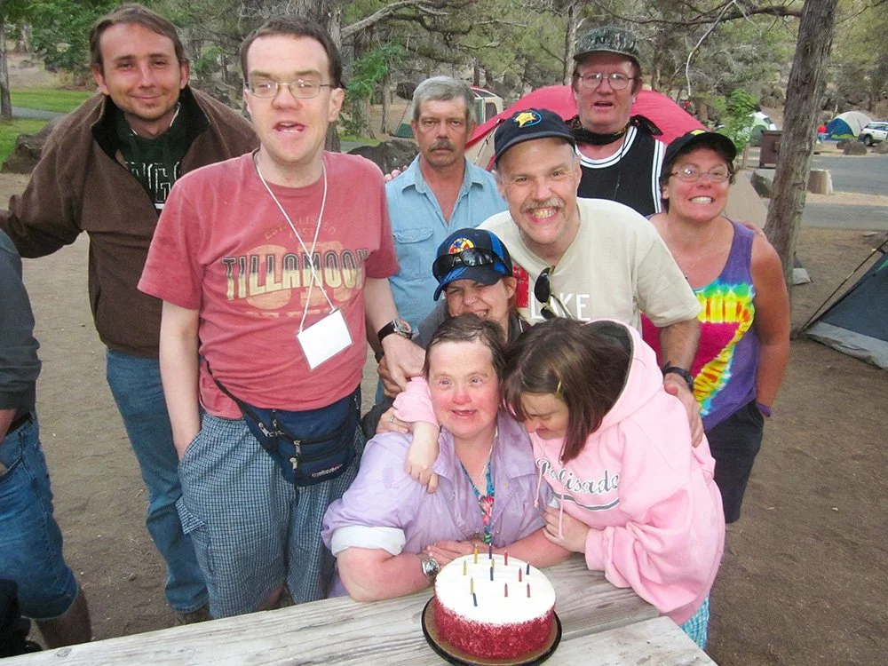 Group of smiling people celebrating a birthday outdoors, gathered around a woman with a birthday cake with lit candles, in a camping area with tents and trees in the background.