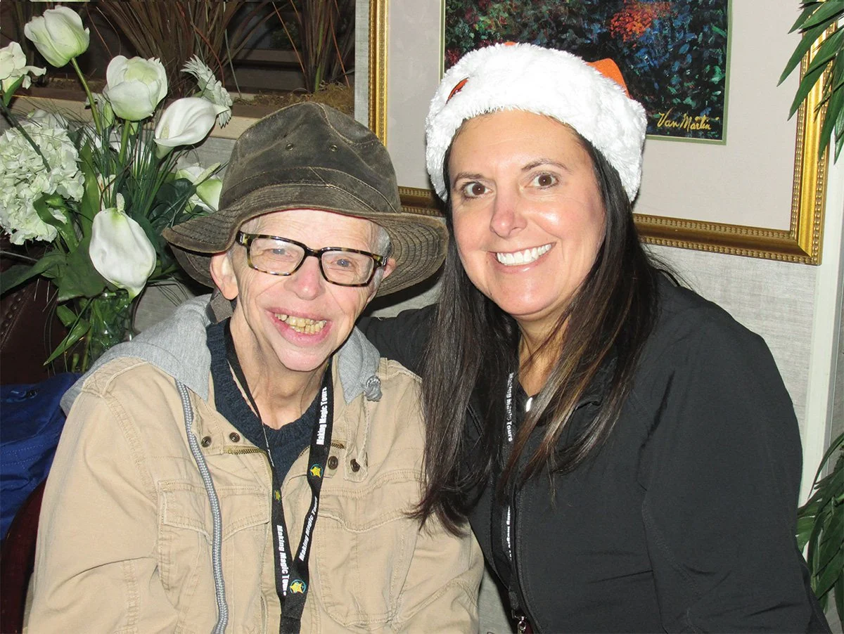 Two women smiling, one wearing a Santa hat and the other wearing a wide-brimmed hat and glasses, sitting indoors with large white flowers and framed artwork in the background.