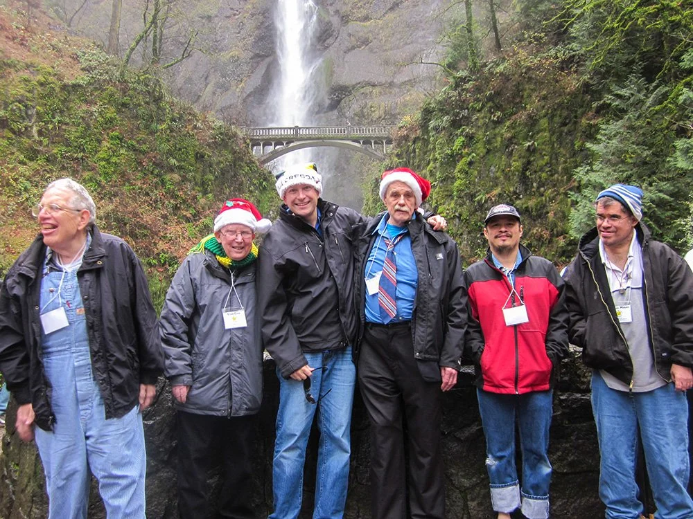 Group of six men wearing winter jackets and Santa hats posing in front of a waterfall with a bridge above it, surrounded by greenery.