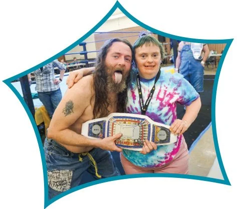 A man with long hair and a beard making a funny face, and a young boy in a tie-dye shirt holding a championship belt, posing together at an event.