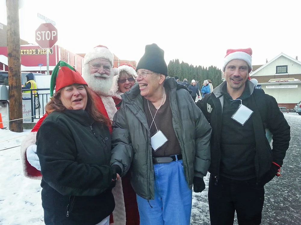 Group of five people dressed in Christmas and winter clothing, standing outdoors on a snowy day, smiling and posing for the photo. Two are wearing Santa hats, one with elf hat, and Santa costume, and one with a black beanie.