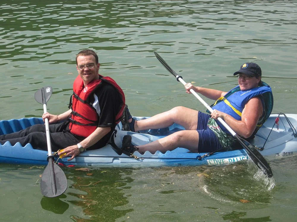 Two men are sitting in a kayak on a calm body of water, smiling and holding paddles. The man in the front is wearing a black shirt and a red life jacket, while the man in the back is wearing a sleeveless shirt, sunglasses, and a blue life jacket.