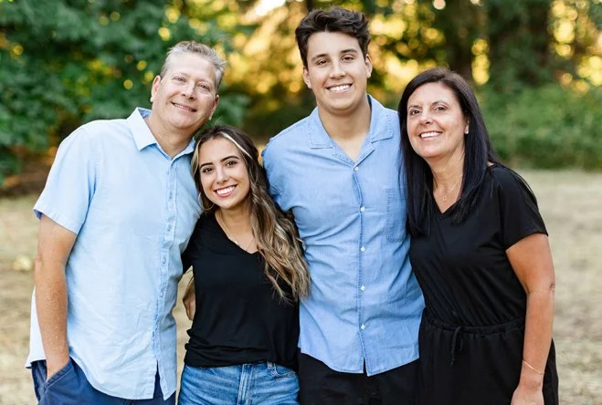 A family of four posing outdoors in a park with trees in the background. The group includes an older man and woman, and a younger man and woman, all smiling.