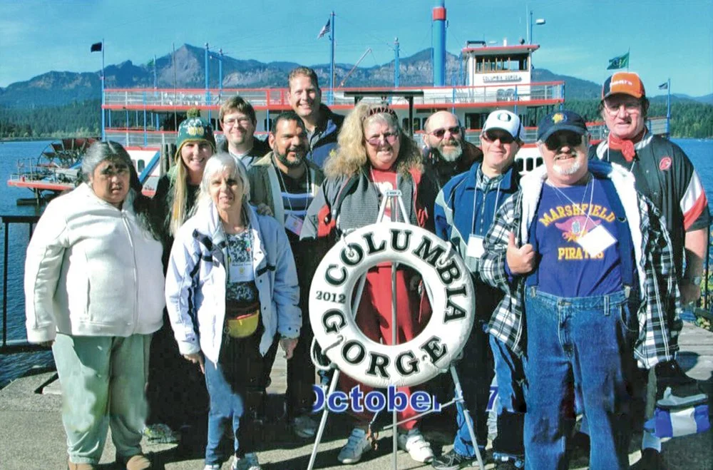 Group of people posing outdoors with a lakeside view, a boat, and mountains in the background, holding a Columbus Gorge sign with October and 2019 on it.