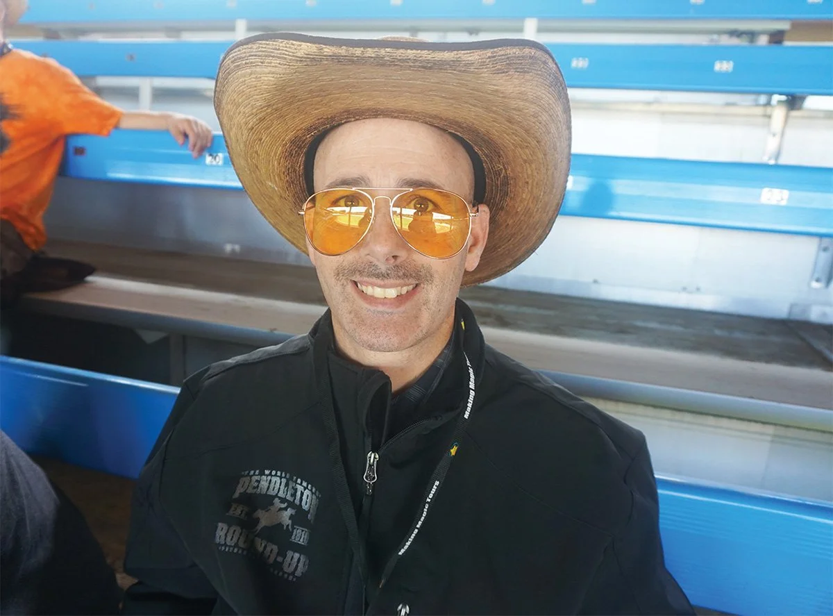 A man wearing a large cowboy hat and orange-tinted sunglasses smiling at the camera, seated on blue stadium seats.