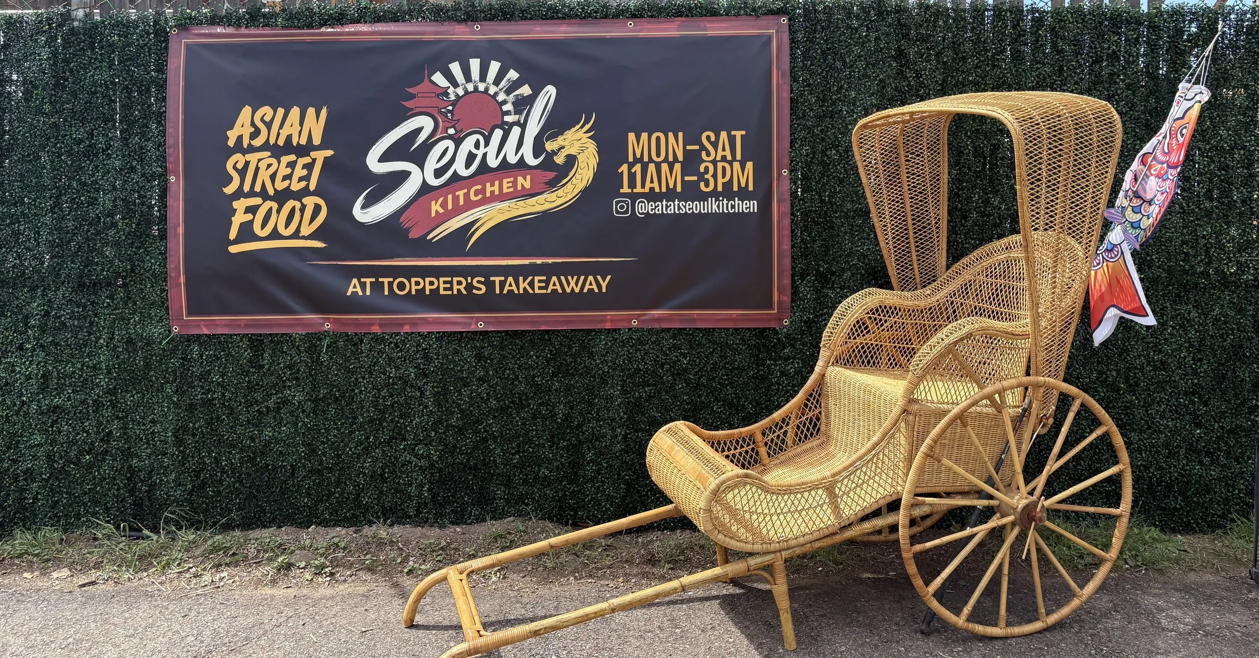 Sign advertising Asian street food at Seoul Kitchen, open Monday through Saturday from 11AM to 3PM, located at Topper's Takeaway, with a rattan chair and parasol standing in front of a green hedge.
