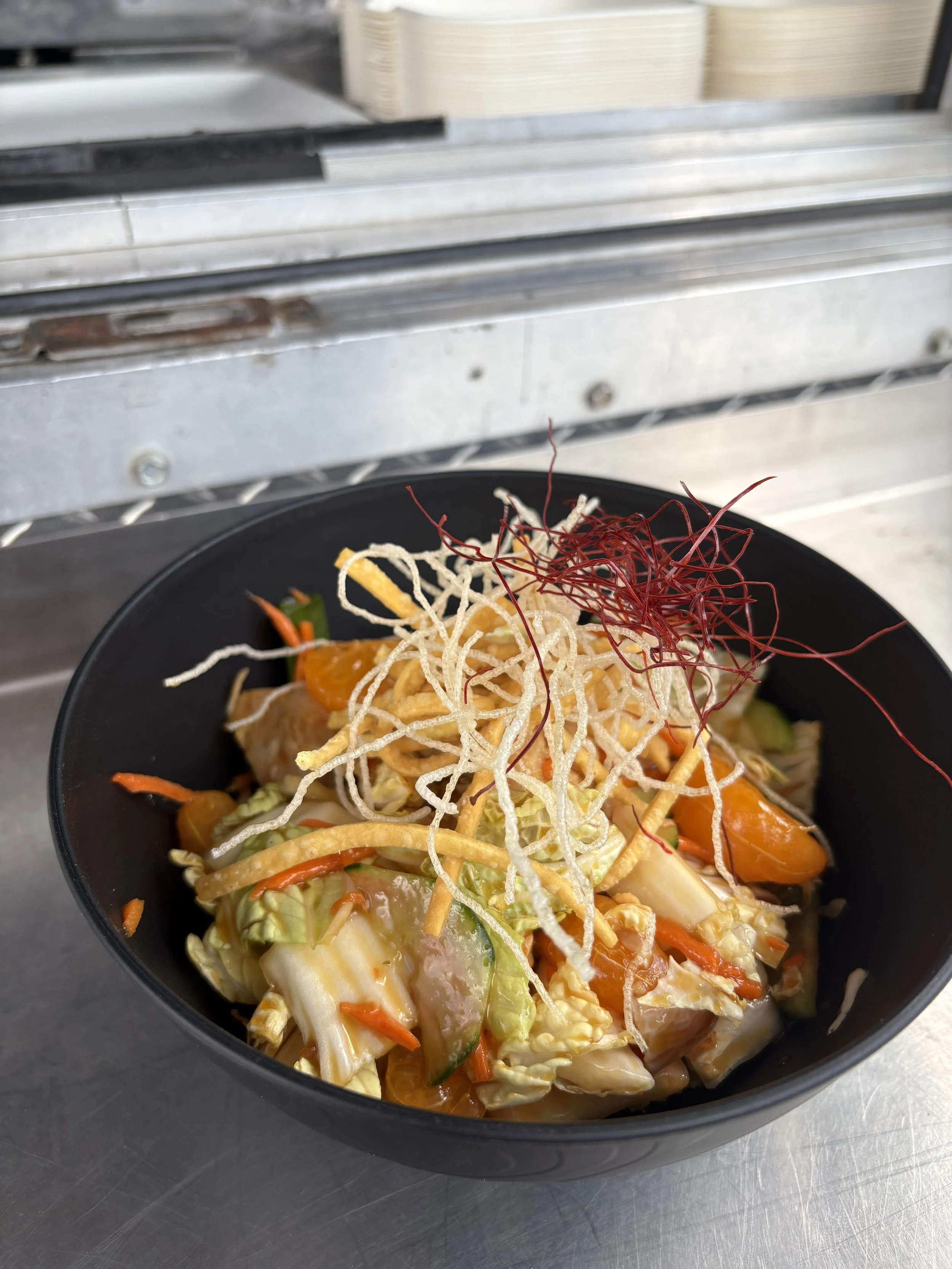 A black bowl of stir-fried vegetables including napa cabbage, carrots, zucchini, and bean sprouts, topped with crispy noodles and red chili threads, placed on a stainless steel counter.