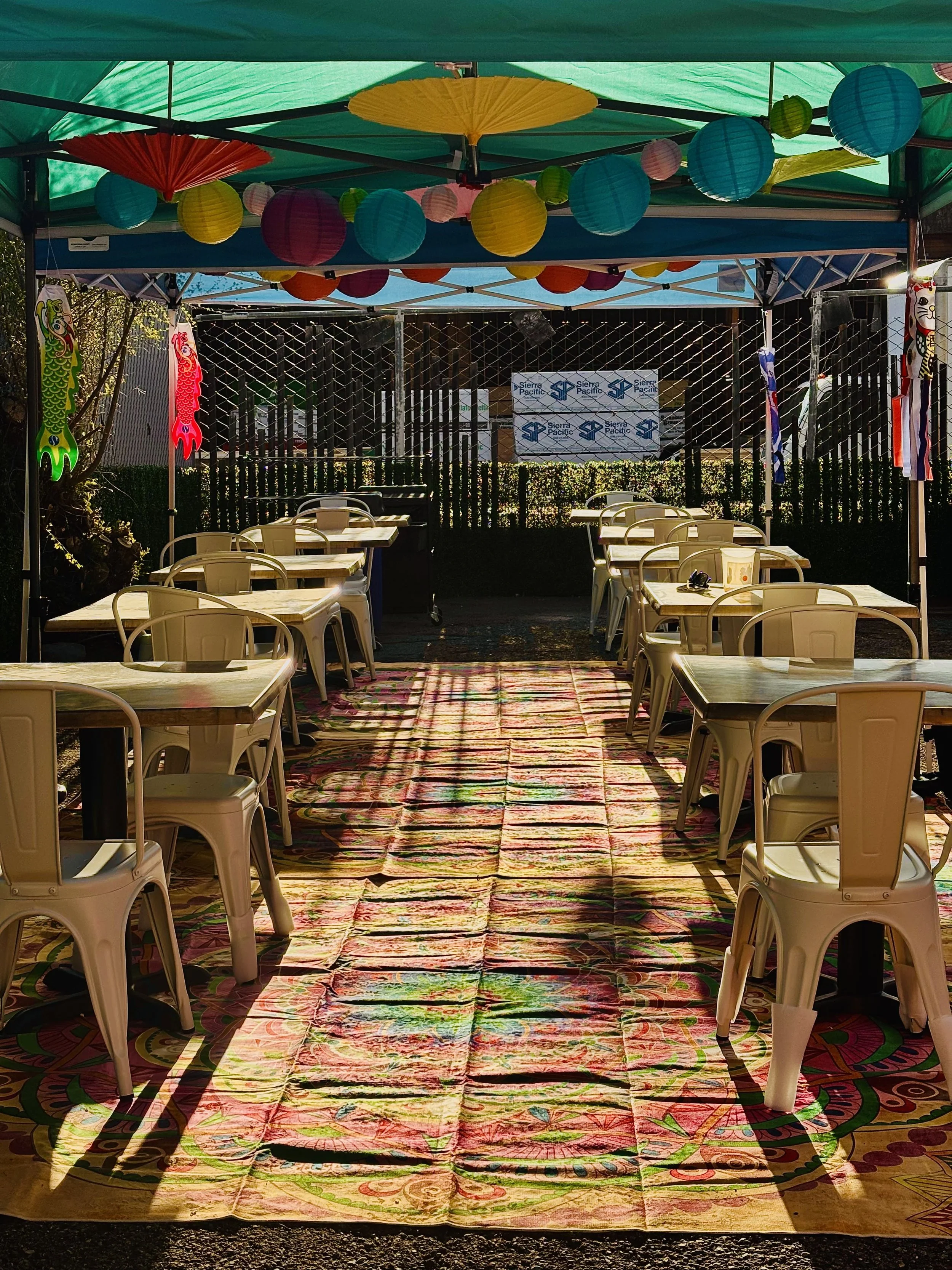 A colorful outdoor seating area with plastic chairs, tables, and a vibrant patterned rug, decorated with paper lanterns and umbrellas under a canopy.