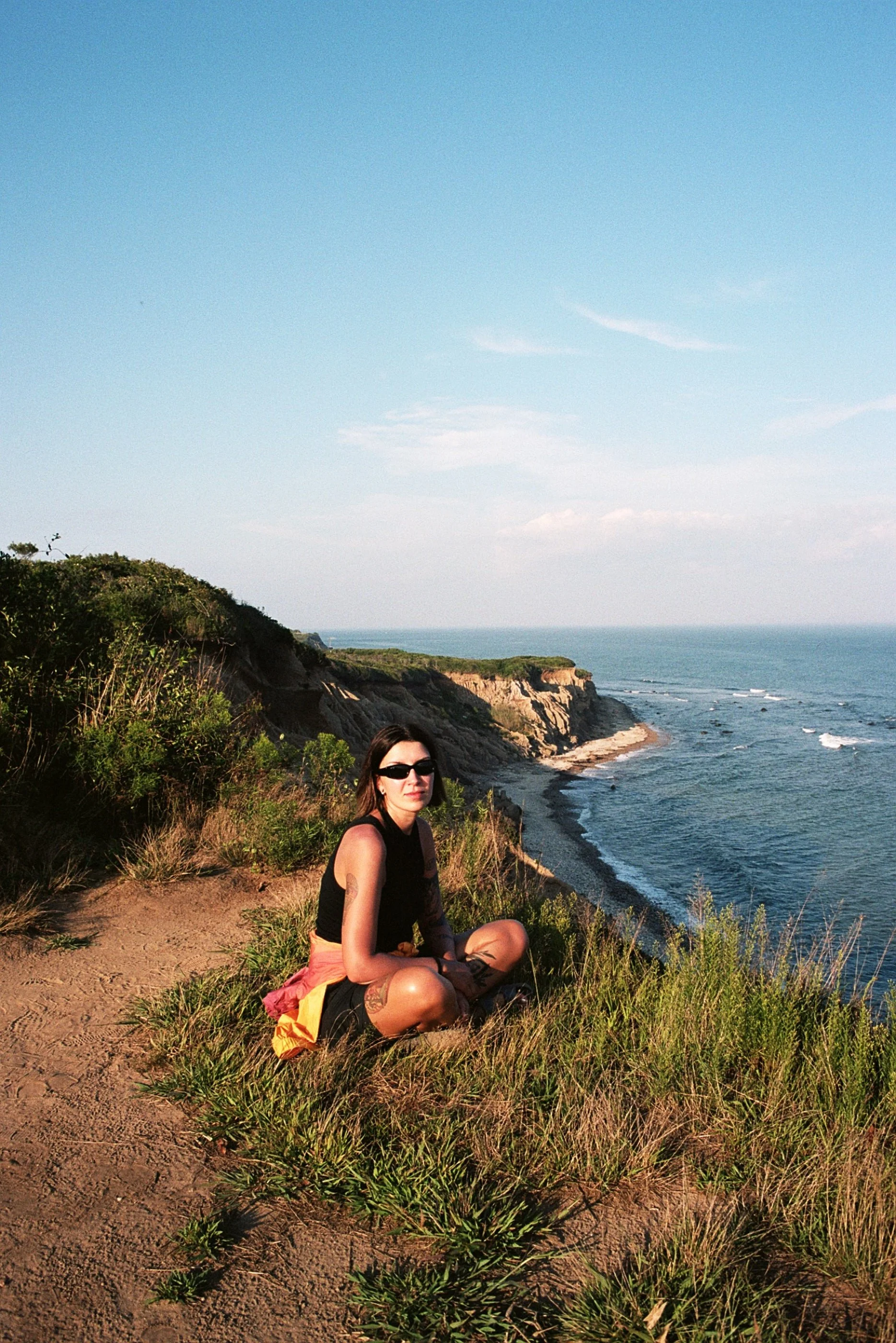 A woman sitting on a grassy cliff overlooking the ocean, wearing sunglasses, a black sleeveless top, and shorts, with coastal cliffs and a sandy beach in the background during daylight.