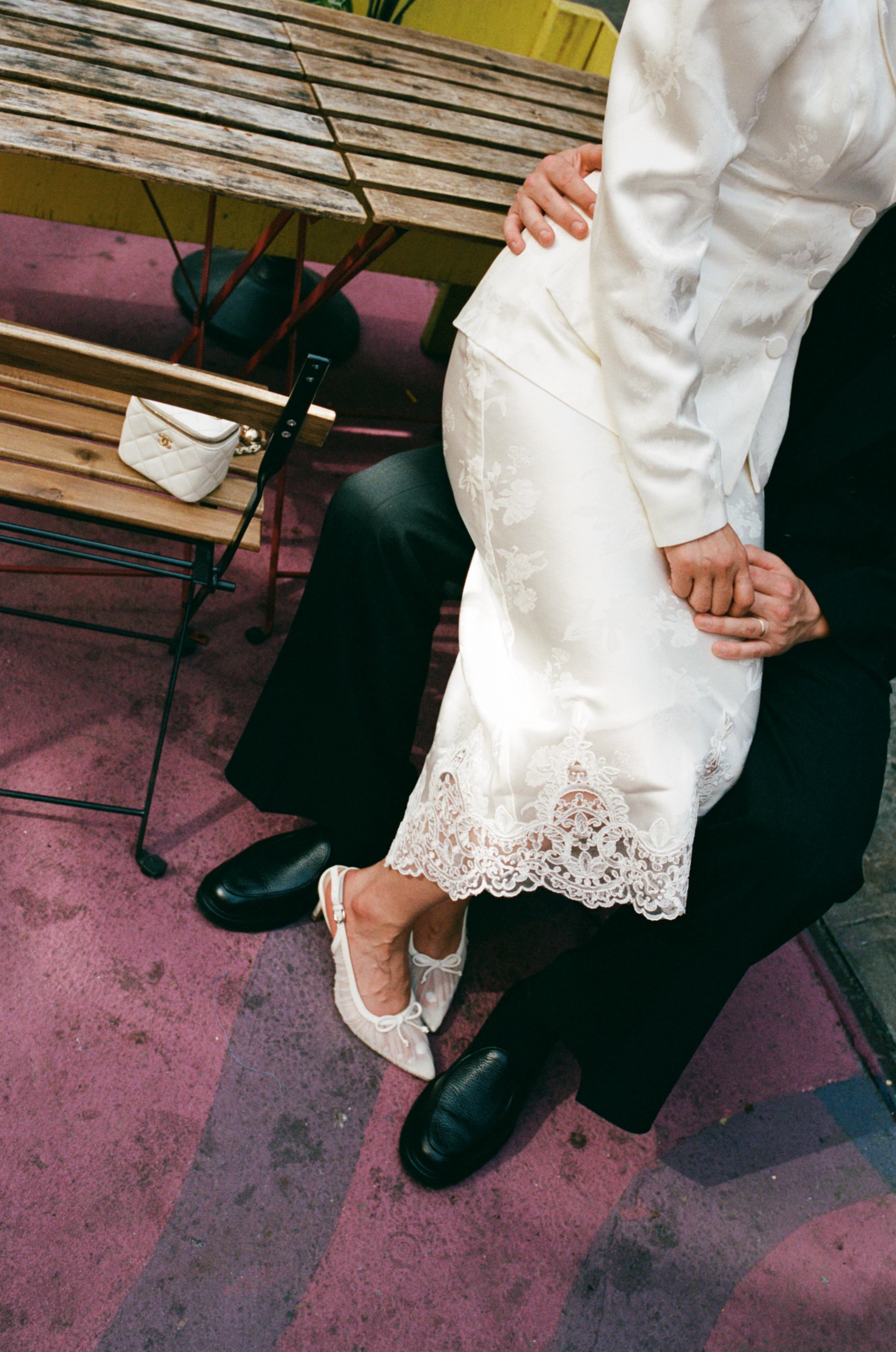 A person dressed in a white, floral lace dress and white shoes is sitting with their knee on someone's lap, their hand resting on their knee, at an outdoor setting with a wooden table, bench, and pink and purple flooring.