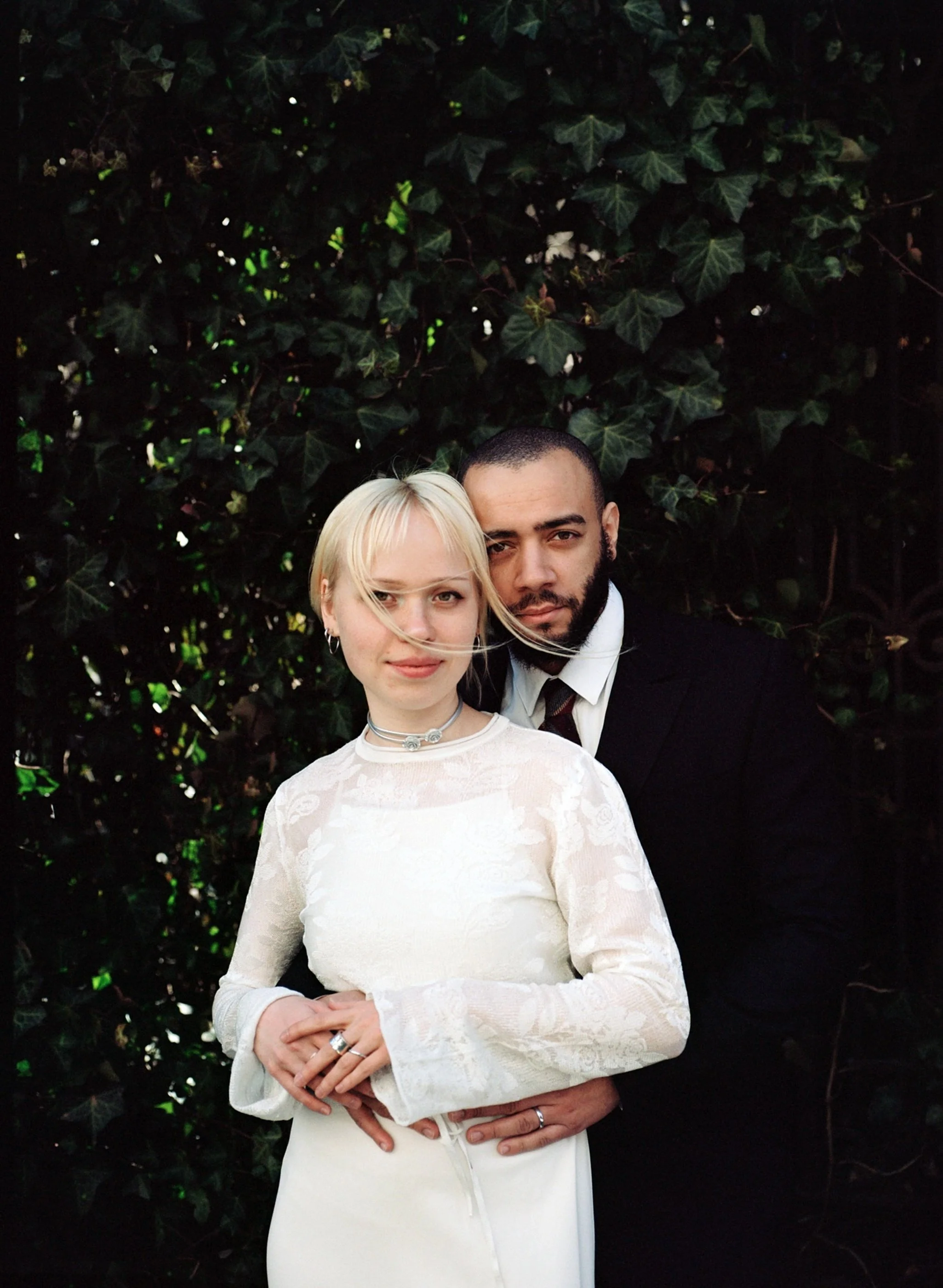 A couple dressed in wedding attire standing in front of dark green leafy background, with the woman's hands held together in front.