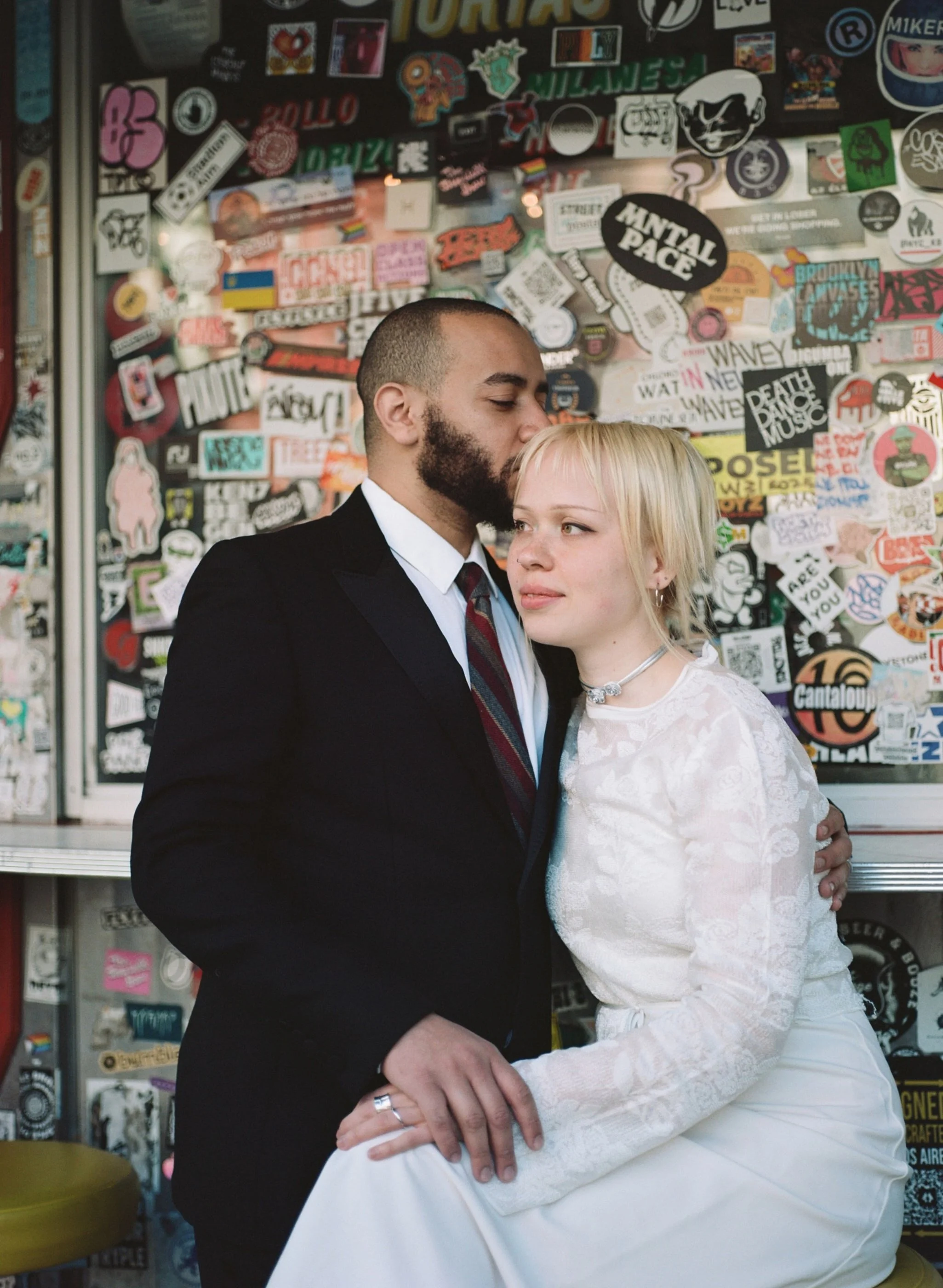 A couple dressed in wedding attire sharing an intimate moment, with the man wearing a black suit and the woman in a white lace dress. They are standing in front of a wall covered with colorful stickers.