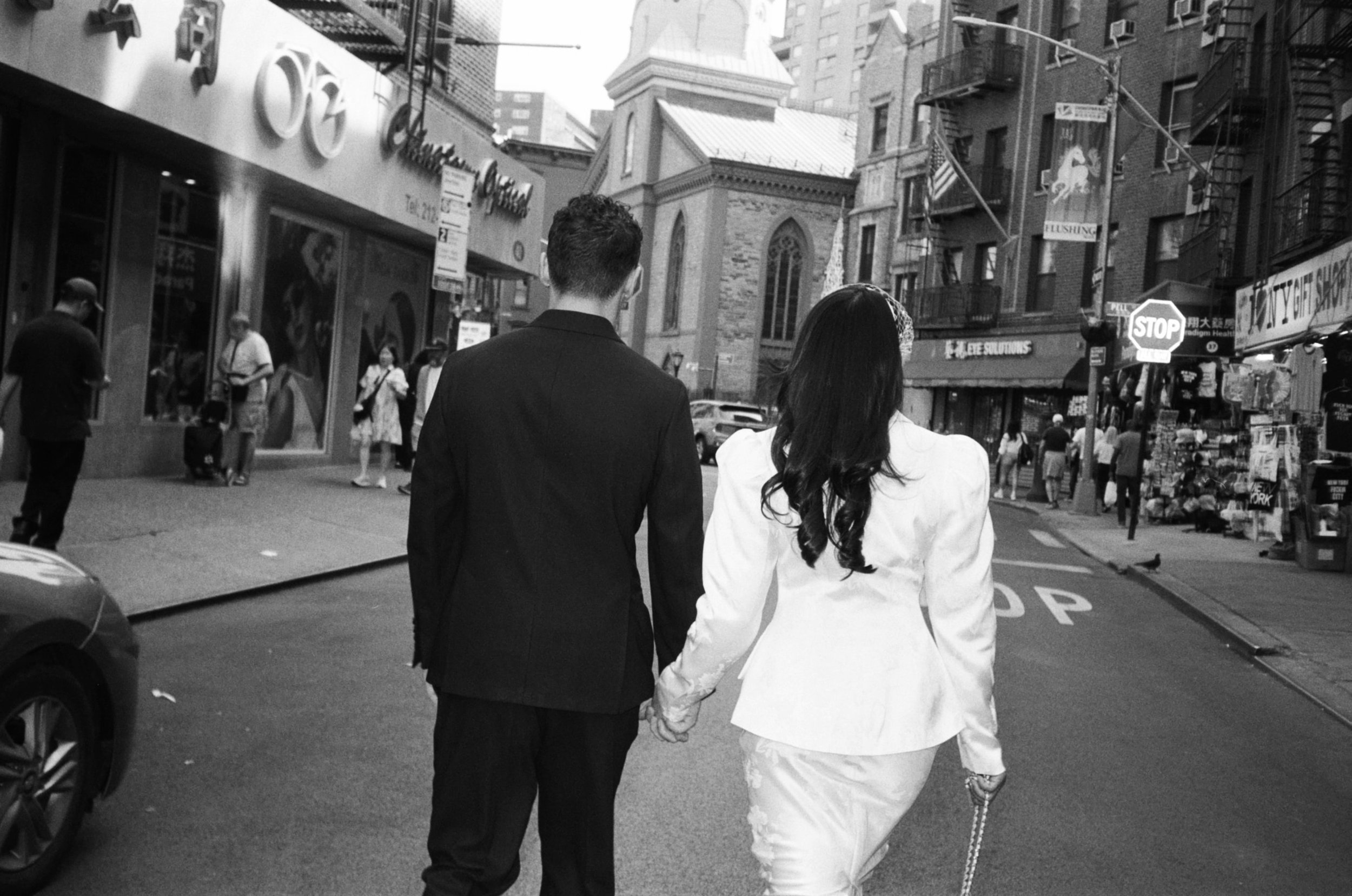 A man and woman holding hands walk down a city street in black and white, surrounded by pedestrians, buildings, and street signs.