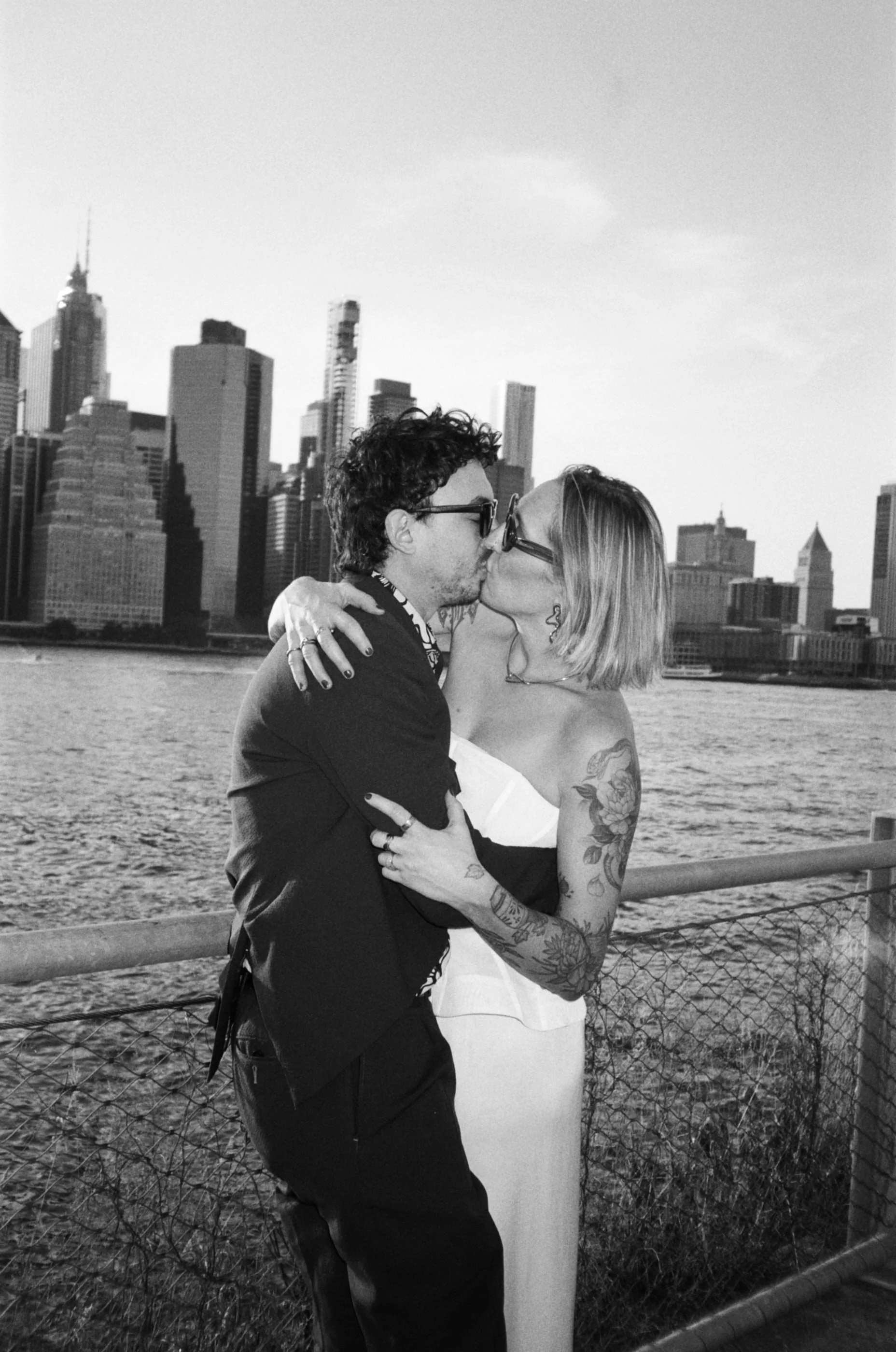 A black and white photo of a couple kissing by a body of water with a city skyline in the background, including tall skyscrapers and the Empire State Building.