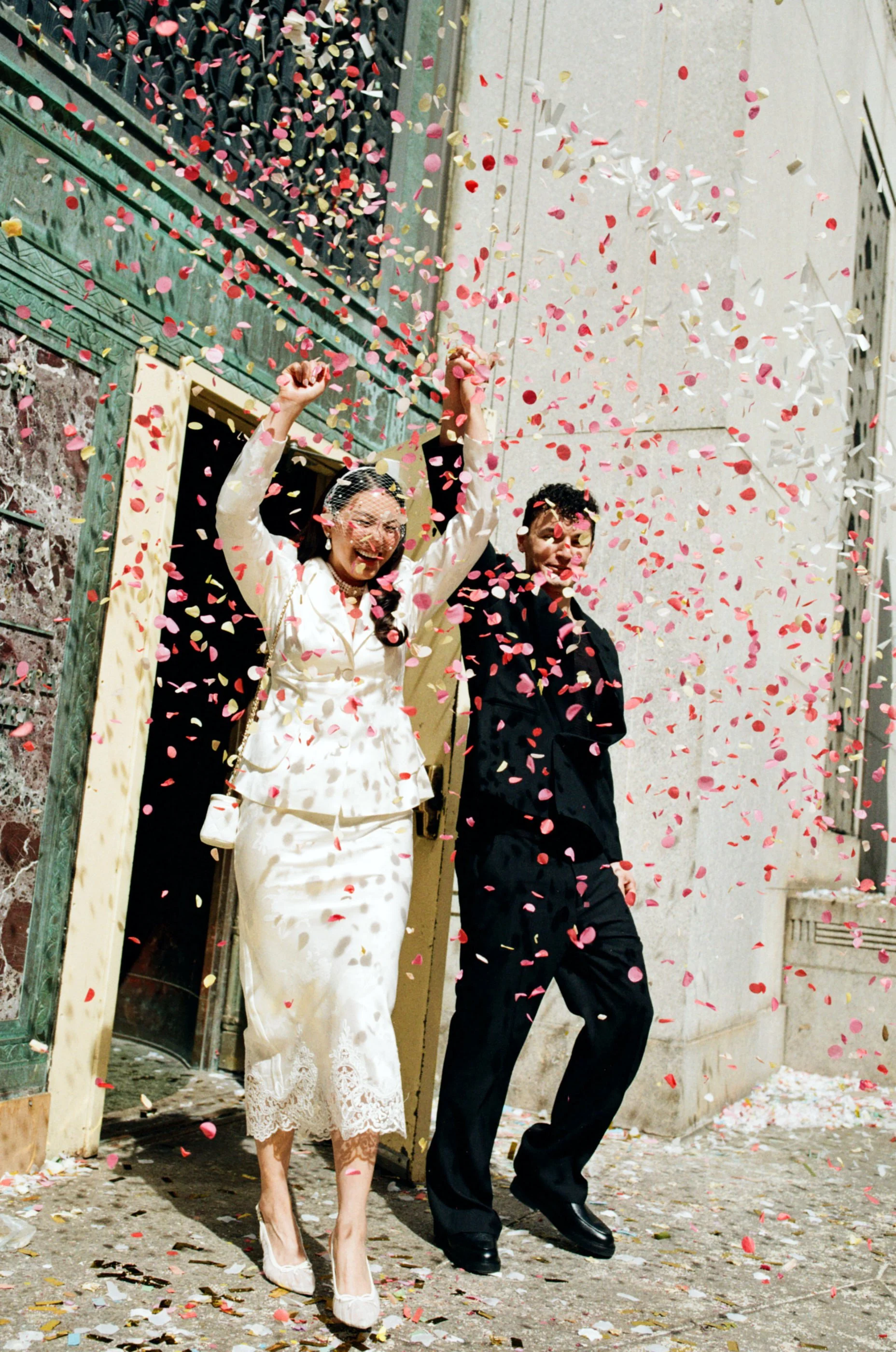 A woman in a white lace dress and a man in a dark suit celebrating under falling confetti on a city street at night.