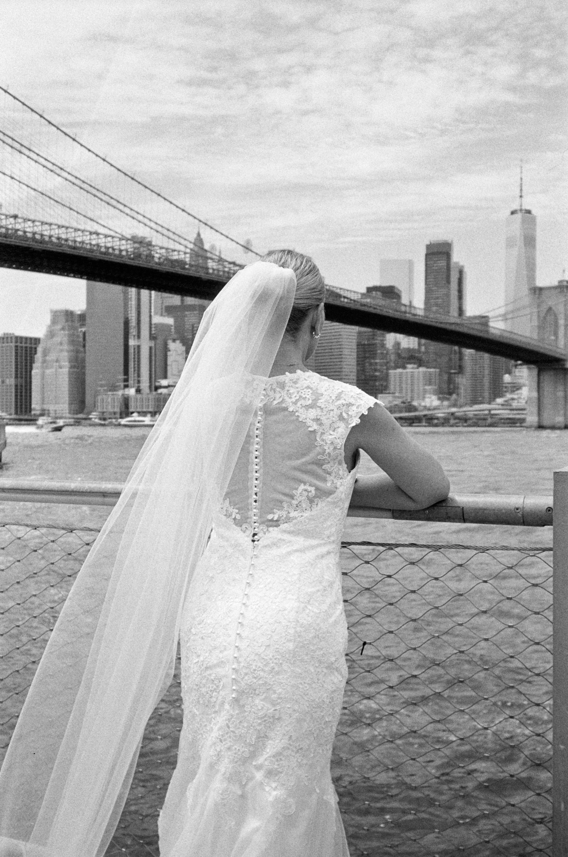 A bride with a veil seen from behind standing by a chain-link fence, overlooking a city skyline featuring the Brooklyn Bridge and tall buildings in black and white.