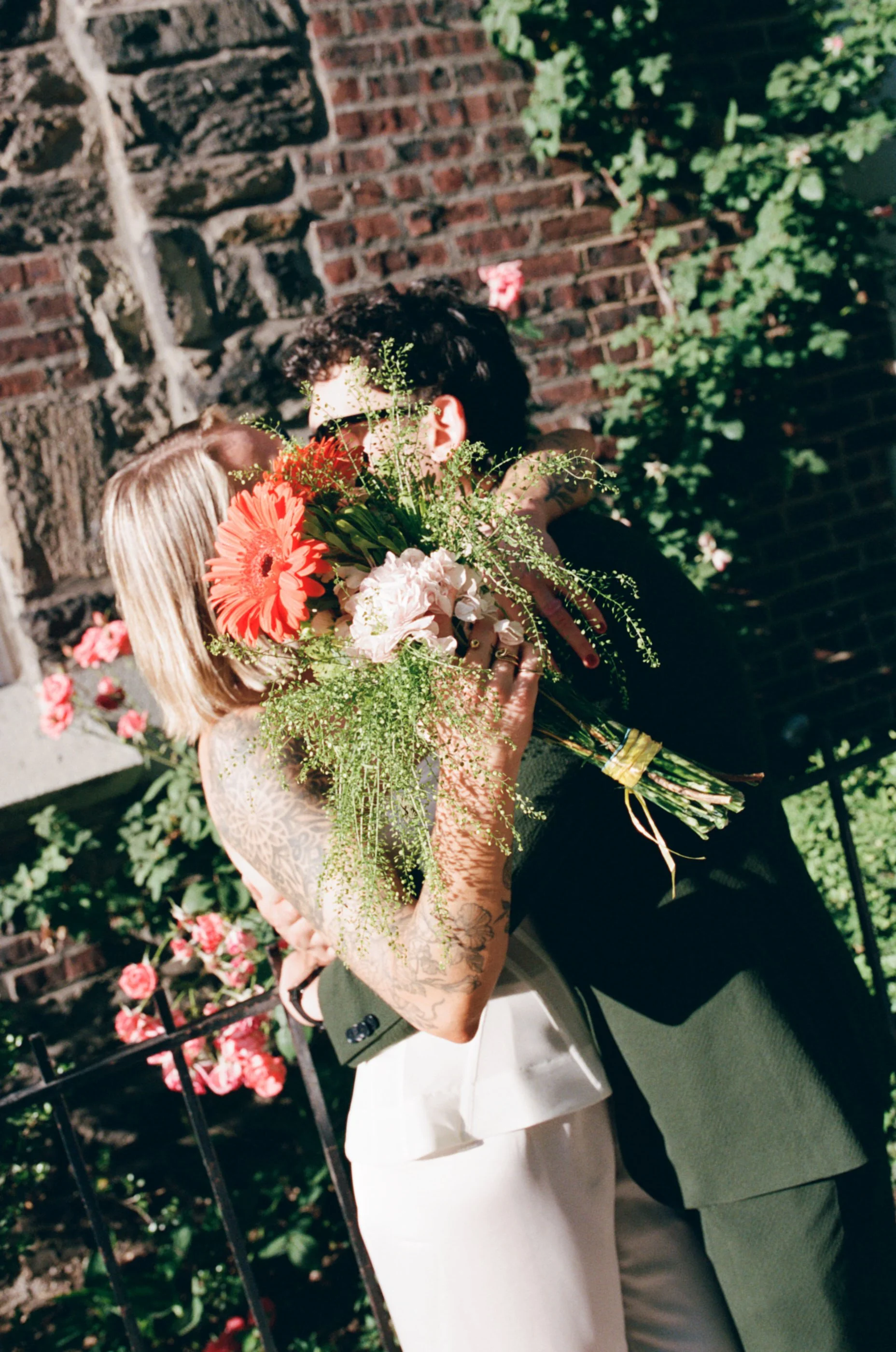 Two people are embracing and holding a bouquet of flowers in front of a brick wall with greenery, during a celebration or wedding.