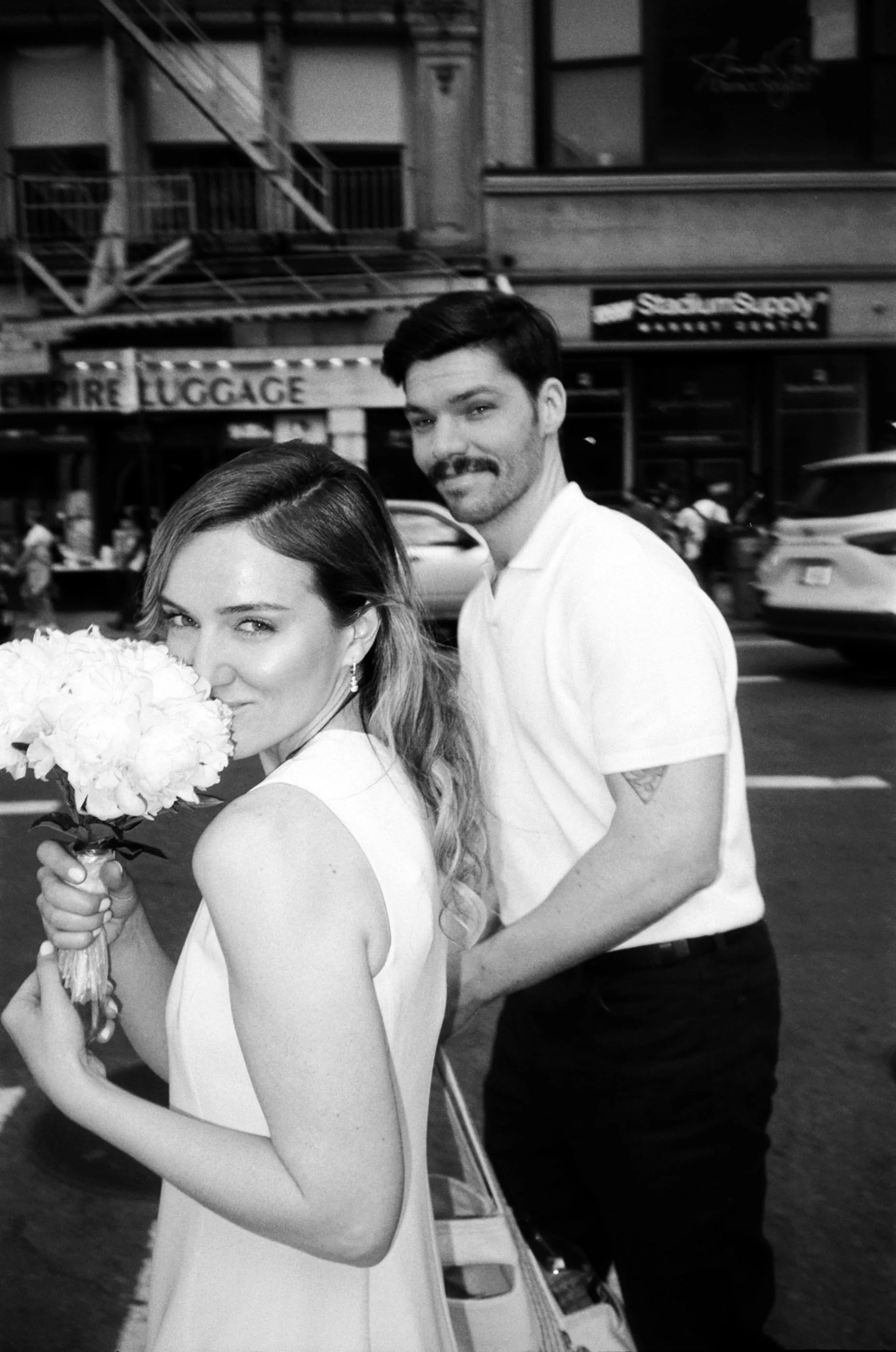 Black and white photo of a woman holding a bouquet of flowers, with a man standing behind her, on a city street with storefronts and cars in the background.