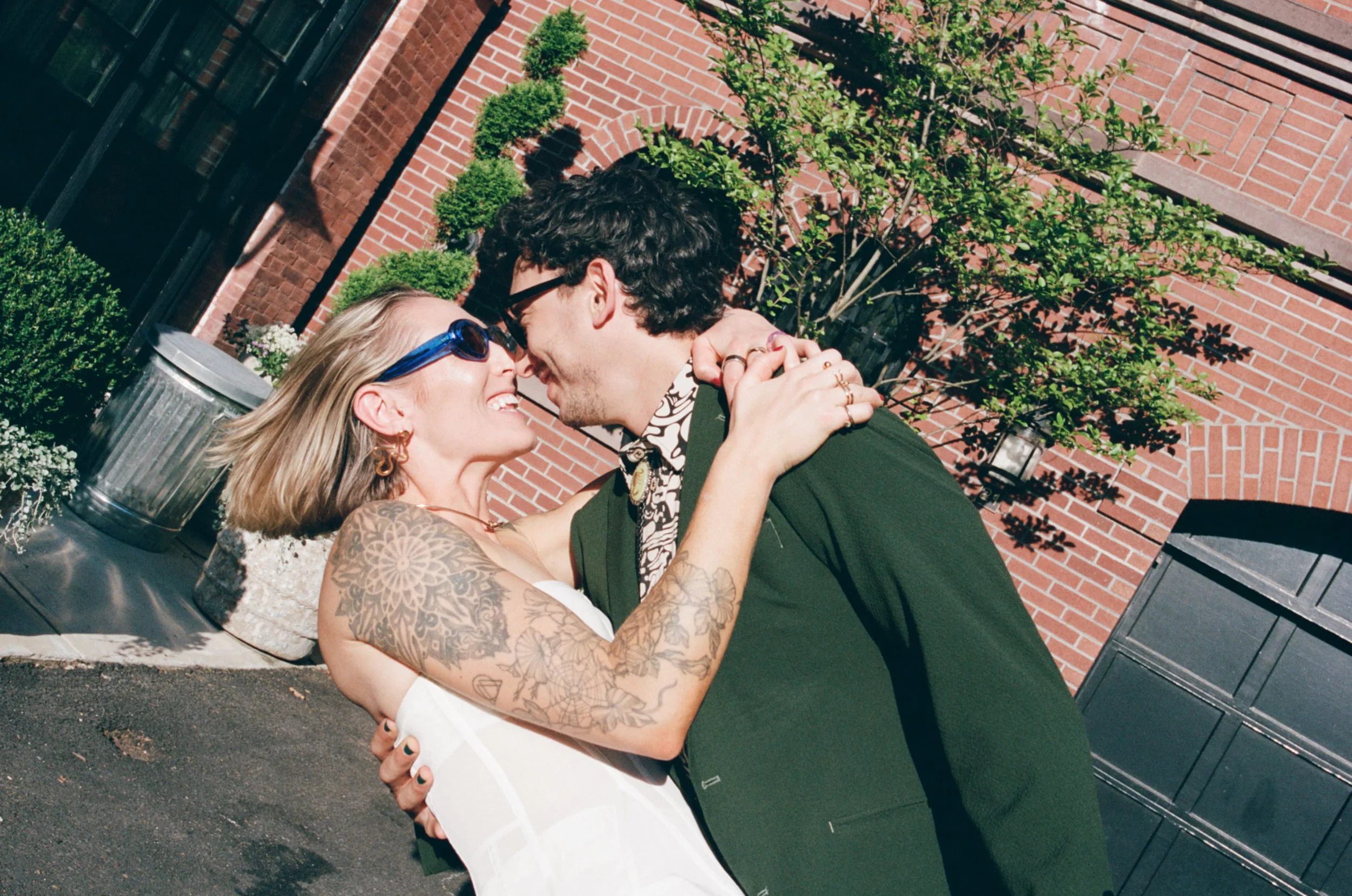 A couple is smiling and leaning their foreheads together outside near a brick building with some greenery, including a small tree and bushes, in the background.