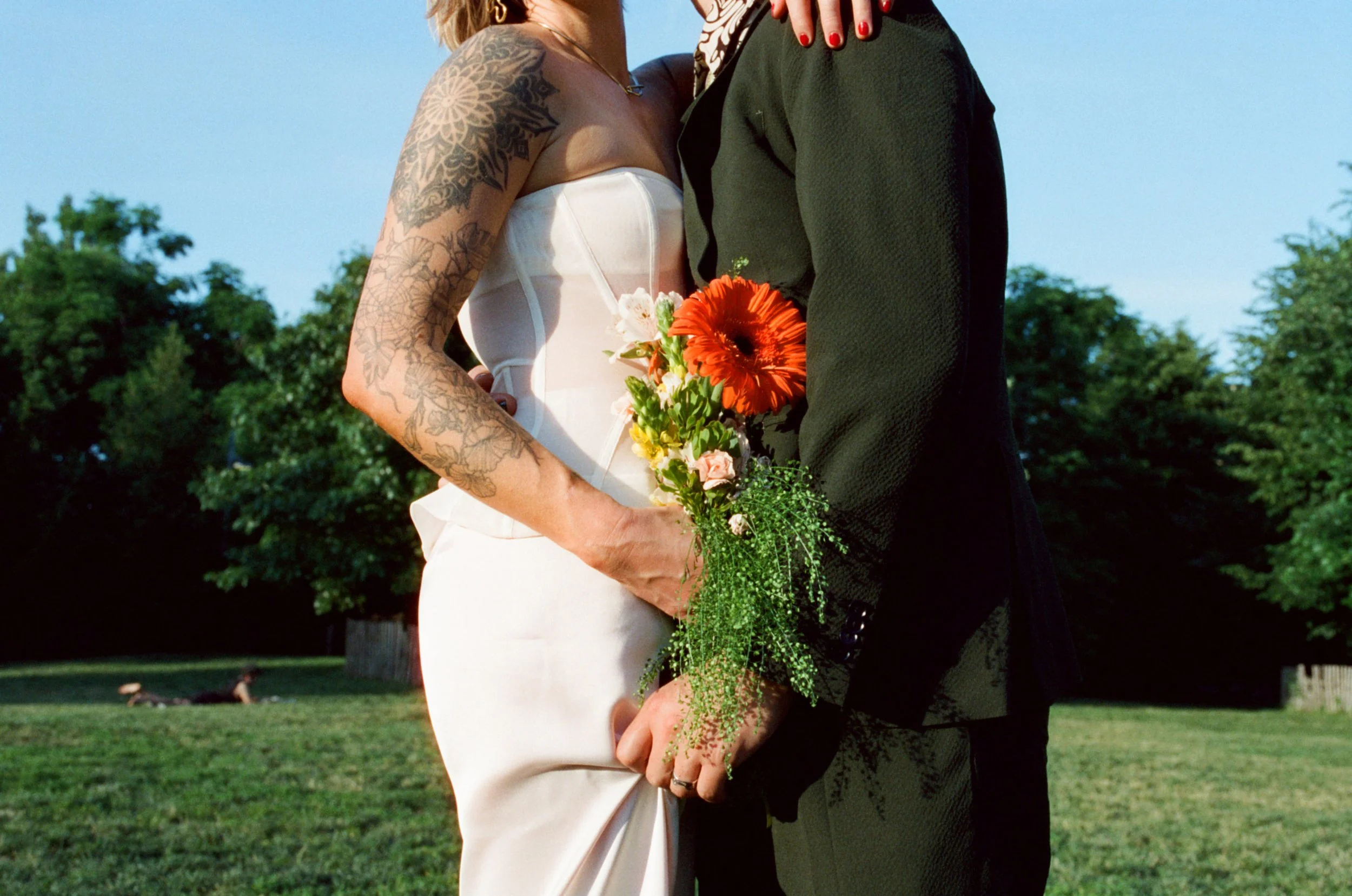 Close-up of a bride and groom standing outdoors with trees in the background. The bride, with visible tattoos on her arm, wears a white strapless wedding dress and holds a bouquet of orange and pink flowers. The groom is dressed in a black suit and tie, with his hand gently holding the bride's hand.