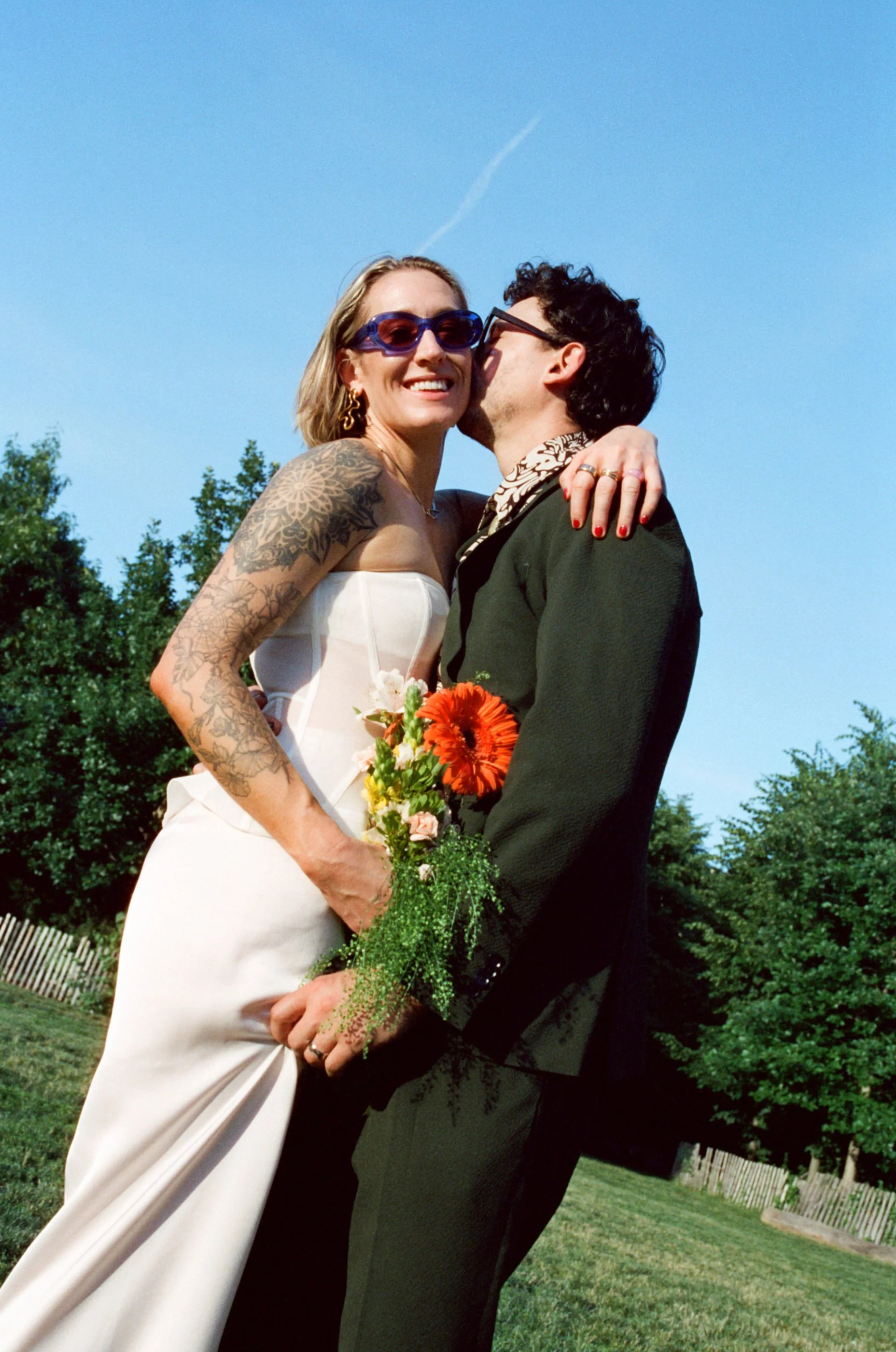 A smiling woman in a white dress and sunglasses shares a kiss with a man in a dark suit and glasses outdoors on a sunny day, holding a bouquet of flowers.