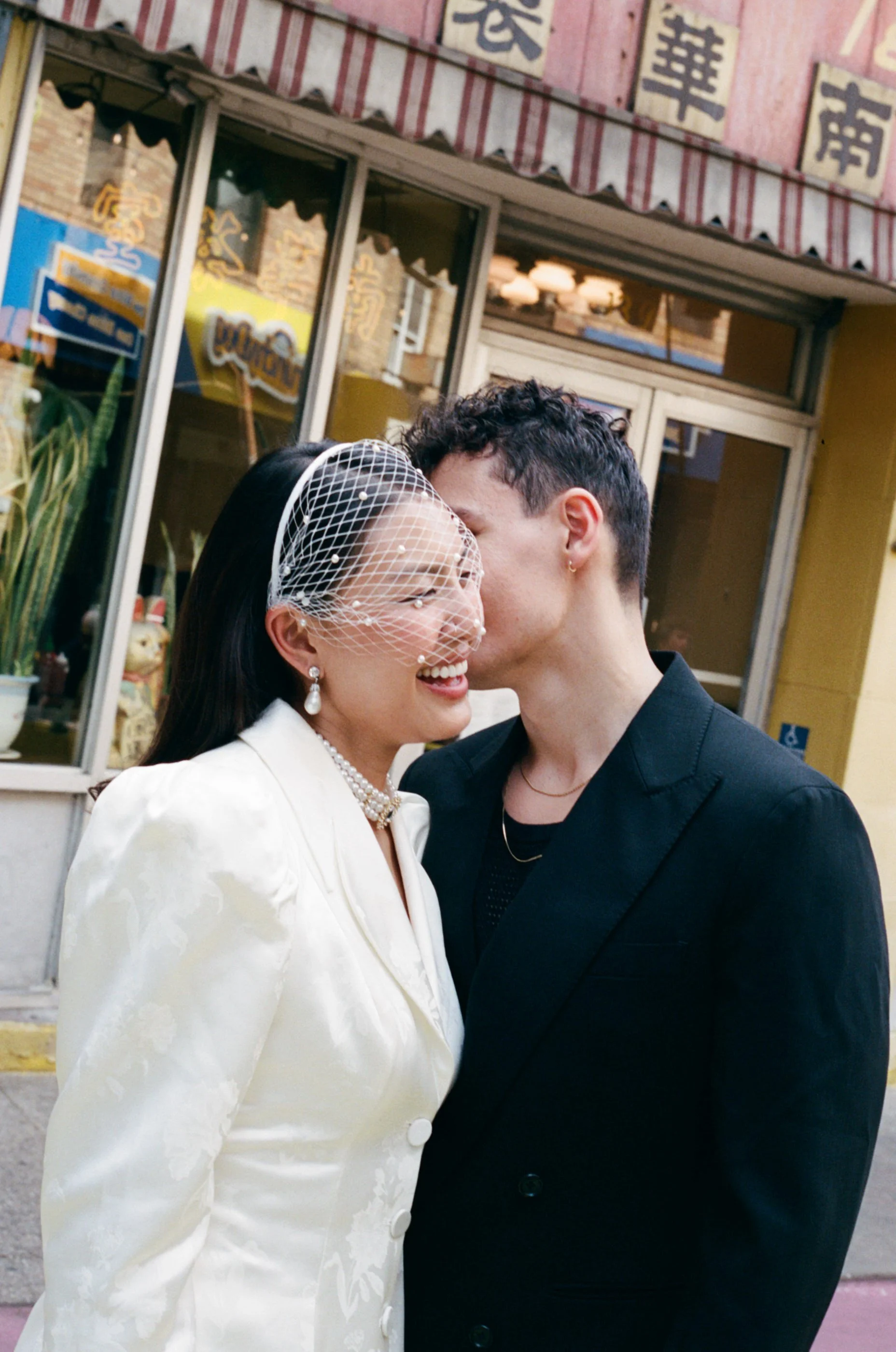 A couple kissing outdoors in front of a building with a sign and storefront windows. The woman is wearing a white dress with a pearl necklace and a netted veil, and the man is in a black suit. The scene appears to be during daytime.