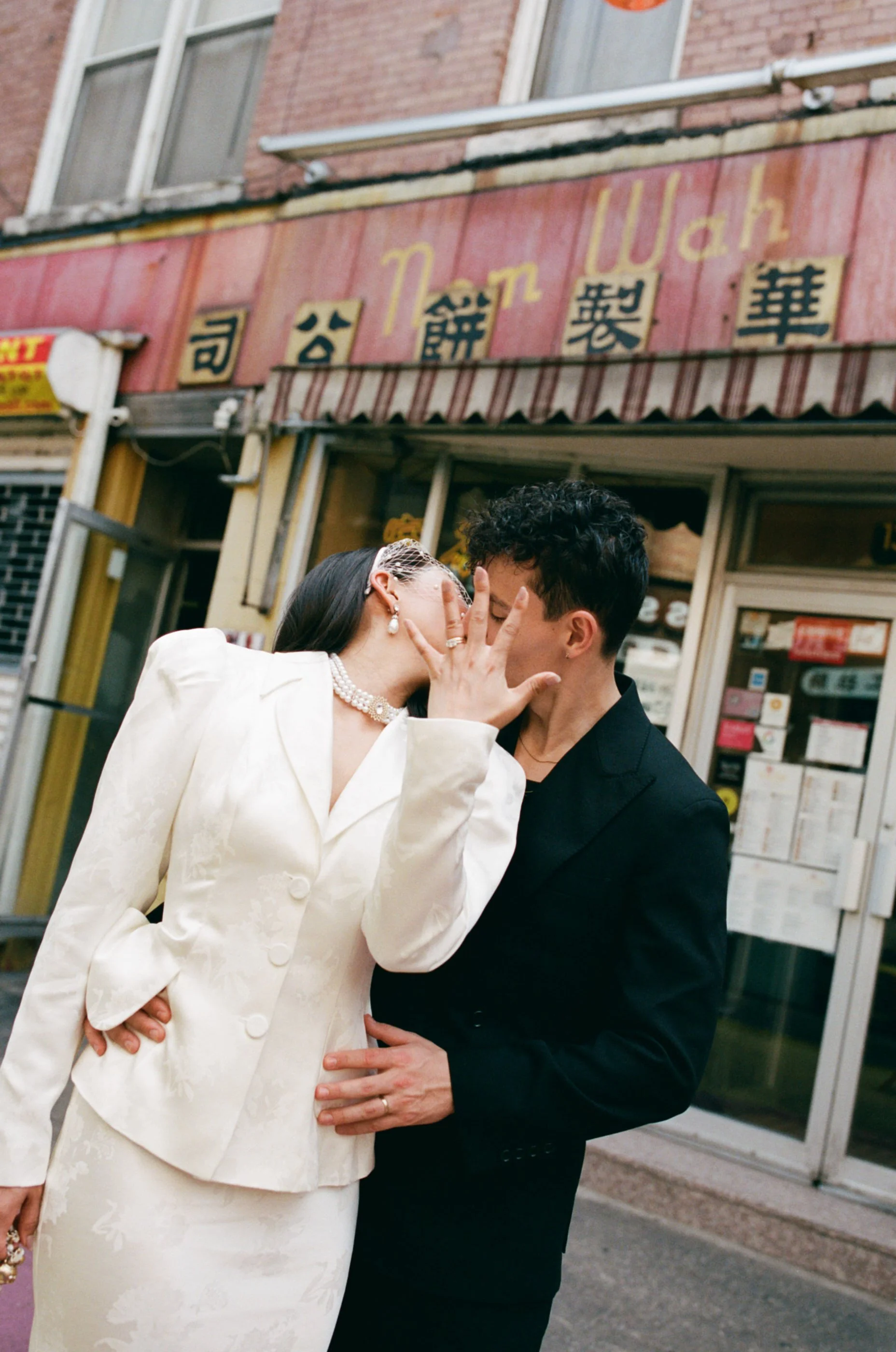 A couple kissing on a city street in front of a Japanese restaurant with a sign in Japanese characters. The woman is dressed in a white blazer and skirt, and the man is wearing a black suit.